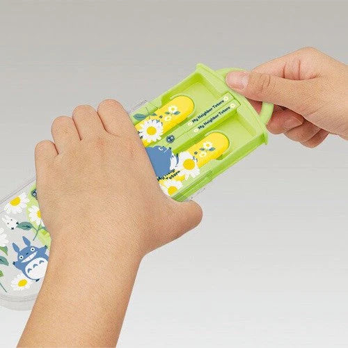 Hands sliding open the green Totoro-themed cutlery case to reveal a fork, spoon, and chopsticks with yellow Totoro-decorated handles.