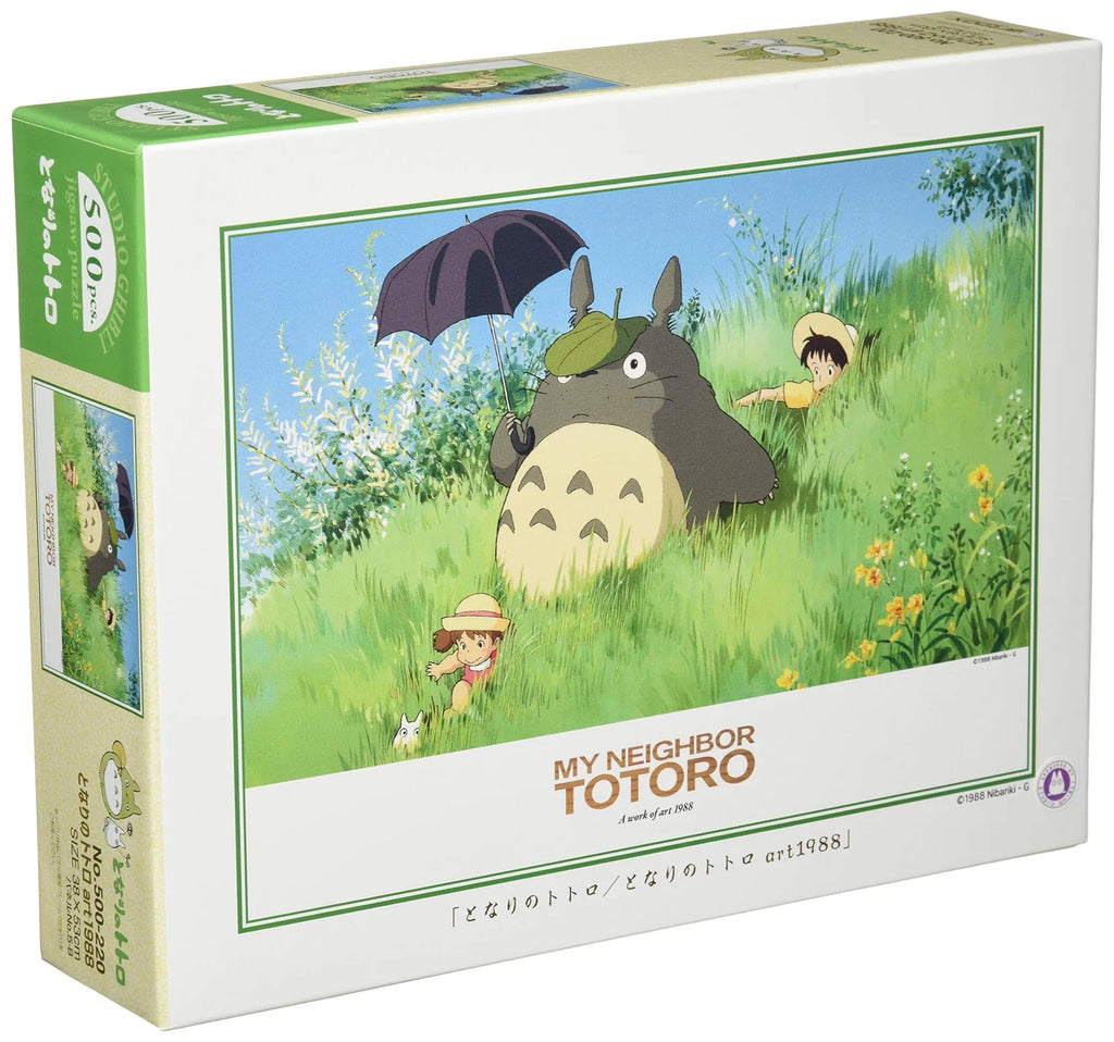 Box of My Neighbor Totoro 500-piece jigsaw puzzle featuring Totoro holding an umbrella, with Satsuki and Mei playing in a grassy meadow on a sunny day.